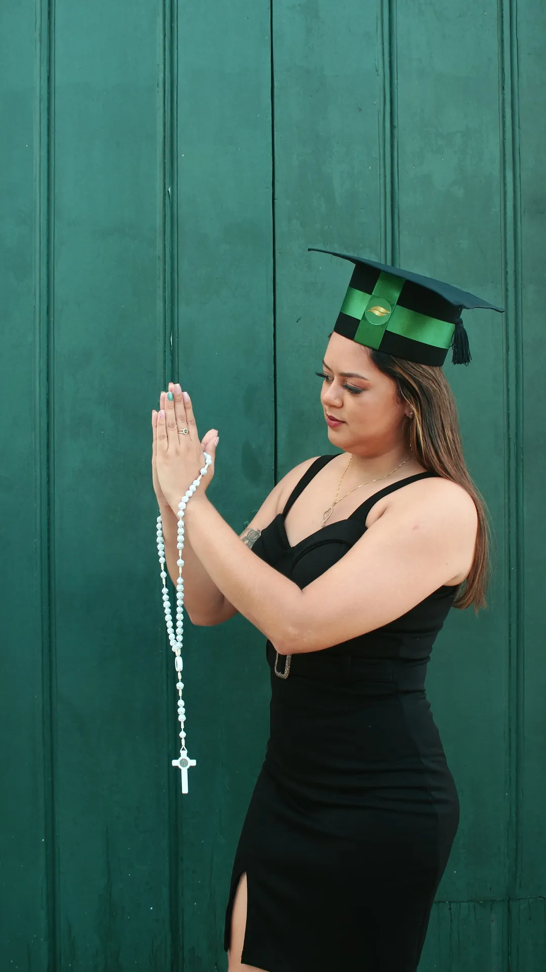 Formanda de vestido preto e capelo verde em prece diante de uma porta verde