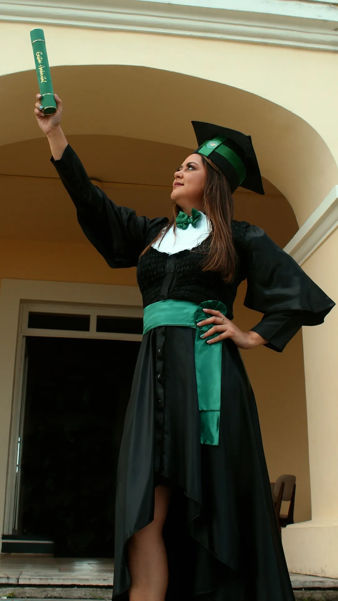 Ensaio de formatura em Belém: formanda de beca e capelo verde erguendo o canudo diante de casarão amarelo