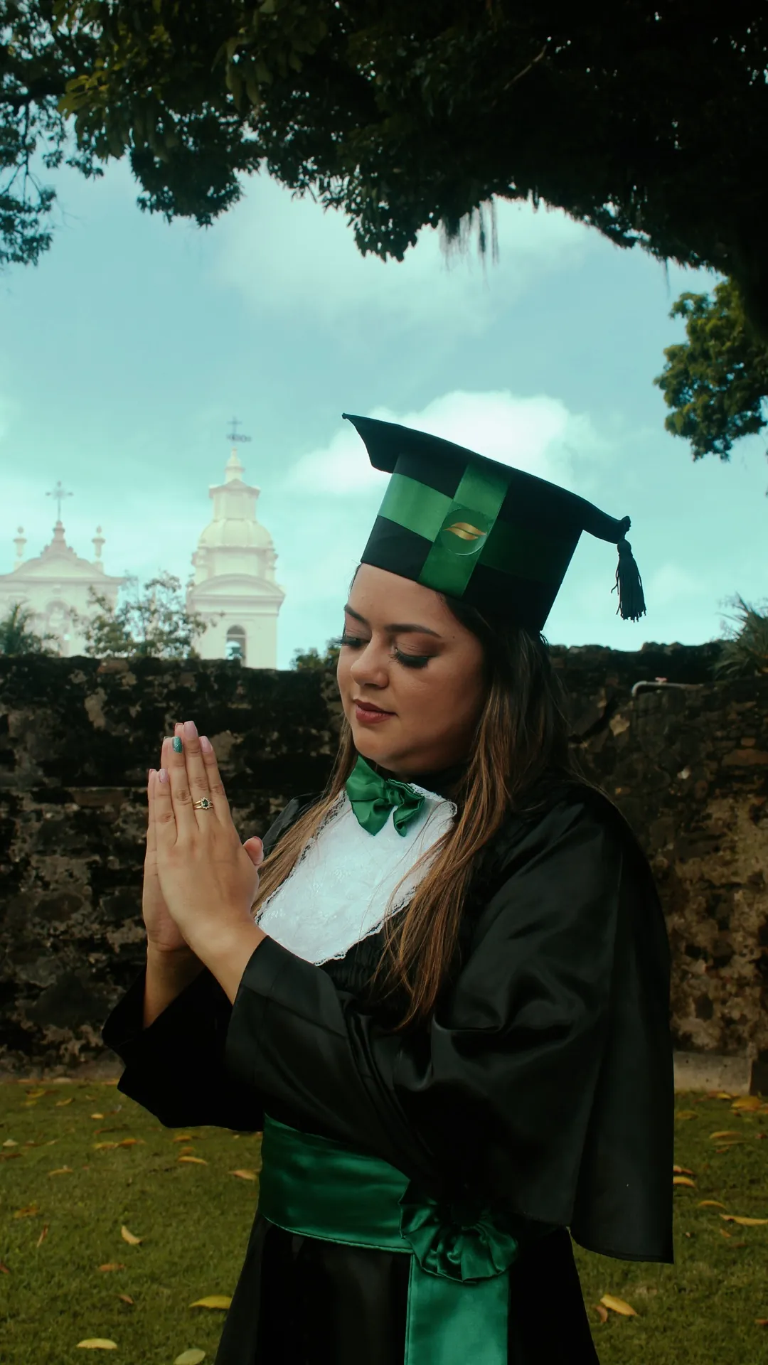 Formanda de capelo verde com as mãos em prece, diante de um muro de pedra