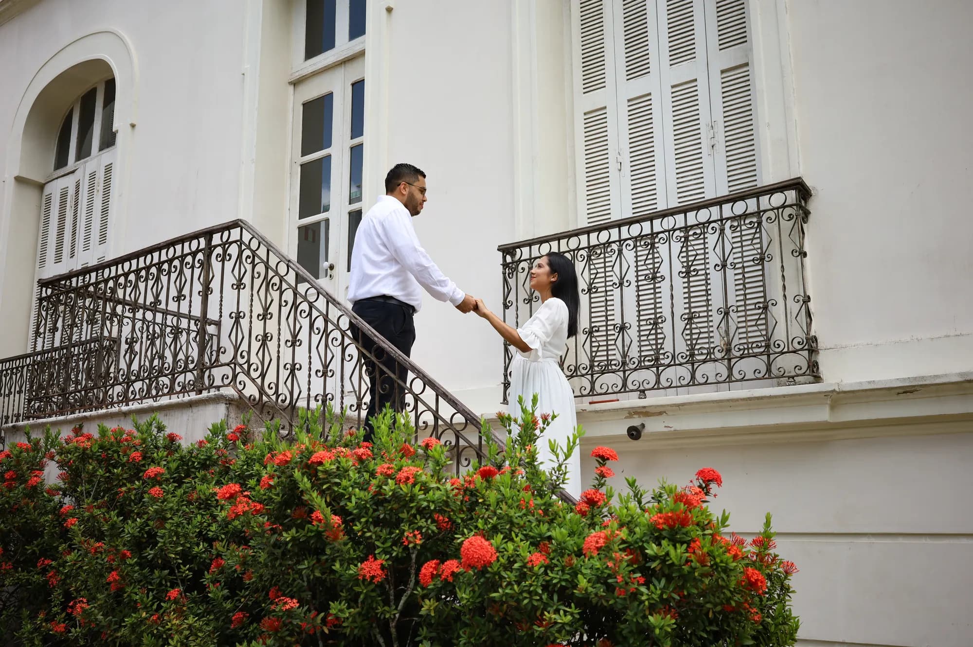 Casal de mãos dadas na sacada de um casarão branco cercado de flores